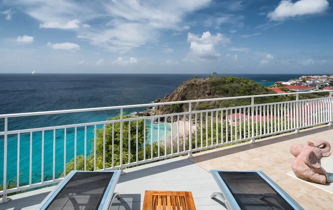 Sleek loungers on warm terracotta tiles face the endless turquoise horizon. A playful pink elephant sculpture adds whimsy while the white railing keeps your view unobstructed—this is where ocean breezes meet island elegance.
