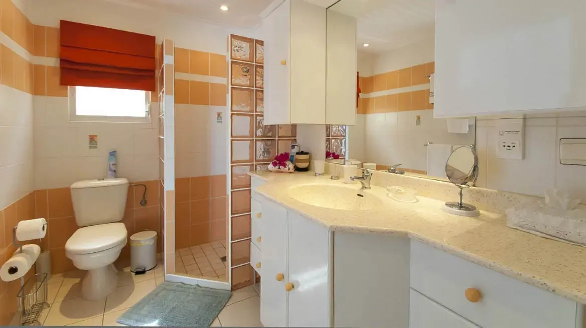 Warm peach-and-white horizontal stripes create a sun-drenched sanctuary. The L-shaped vanity with expansive countertop offers abundant space, while a glass block partition adds modern elegance to the walk-in shower.