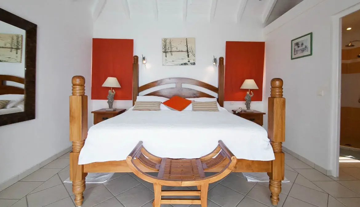 A handcrafted four-poster king bed anchors this bright retreat, dressed in crisp white with vibrant orange accents. Curved wooden bench, vaulted ceilings, and artisanal lighting create a warm Caribbean sanctuary where mornings start with soft natural light.