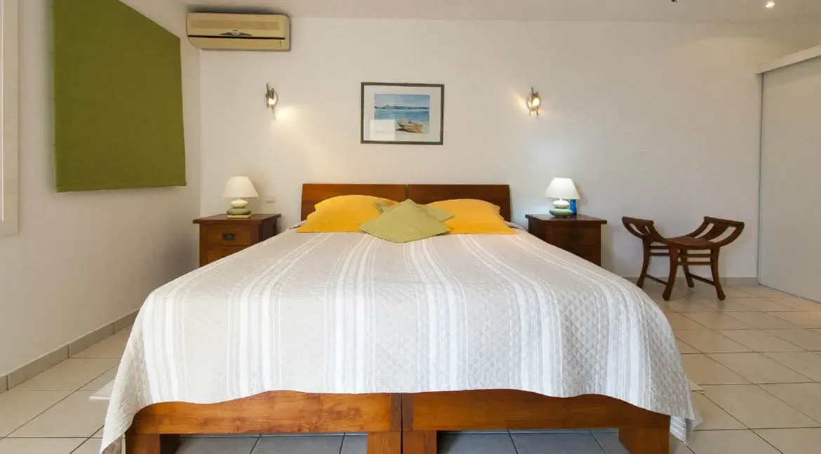 A handcrafted wooden platform bed anchors this sun-drenched retreat, dressed in crisp linens with vibrant canary-yellow pillows. Starburst wall sconces and coastal artwork create an elegant sanctuary, while sliding doors reveal ample closet space for extended stays.