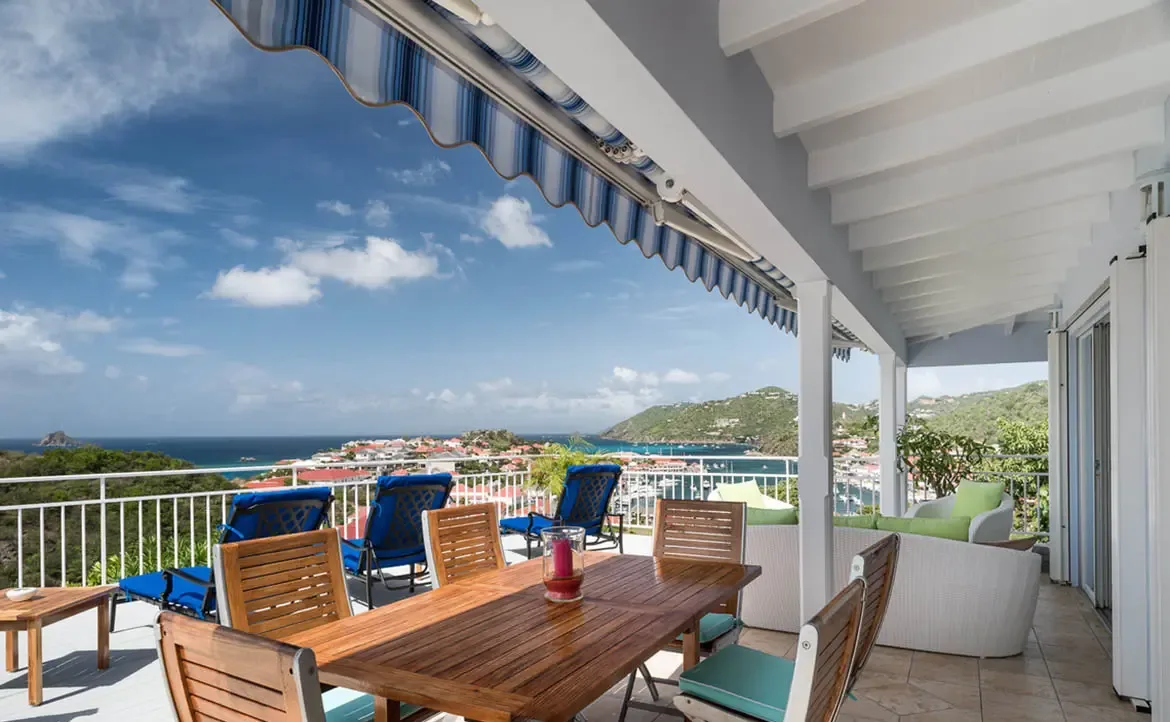 The covered terrace centers around a solid wood dining table for six, with a curved white wicker sectional and lime-green cushions inviting sunset gatherings. Cobalt-blue loungers complete this indoor-outdoor haven overlooking Gustavia's iconic red rooftops.