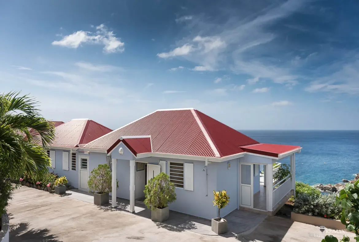 The iconic red corrugated roof crowns this classic West Indian villa, where soft blue walls and white trim echo the Caribbean sky. A white-railed veranda extends toward the endless ocean—authentic island architecture meets panoramic serenity.