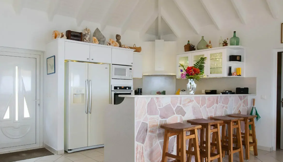 A stone-clad breakfast bar topped with fresh tropical flowers anchors this inviting kitchen. Rustic barstools, white cabinetry, and oversized conch shells create the perfect gathering spot for morning coffee or evening cocktails.