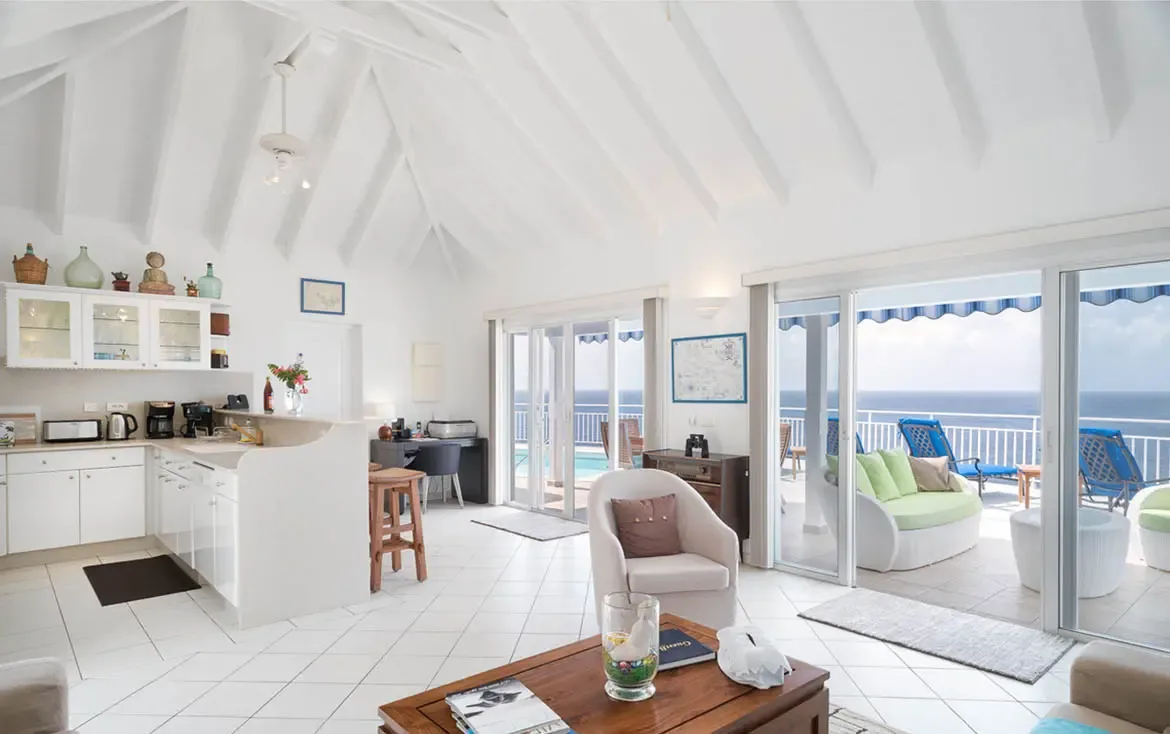 Floor-to-ceiling glass doors frame the turquoise ocean beyond, merging indoor comfort with Caribbean beauty. The all-white kitchen and cozy living area share the same stunning vista—where cooking, dining, and relaxing all happen with the sea in sight.