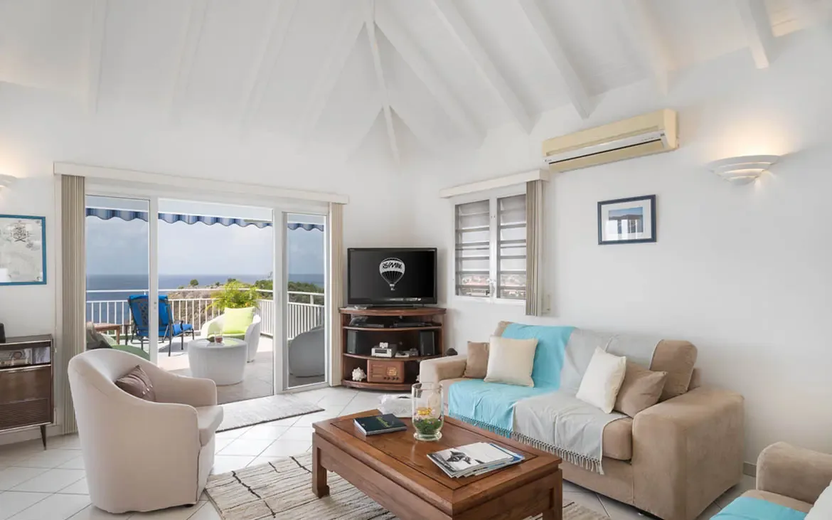 Soaring white vaulted ceilings crown this airy living space, where a tan sofa and plush armchair invite leisurely afternoons. Floor-to-ceiling glass doors vanish to merge indoor comfort with the terrace and that endless ocean view.
