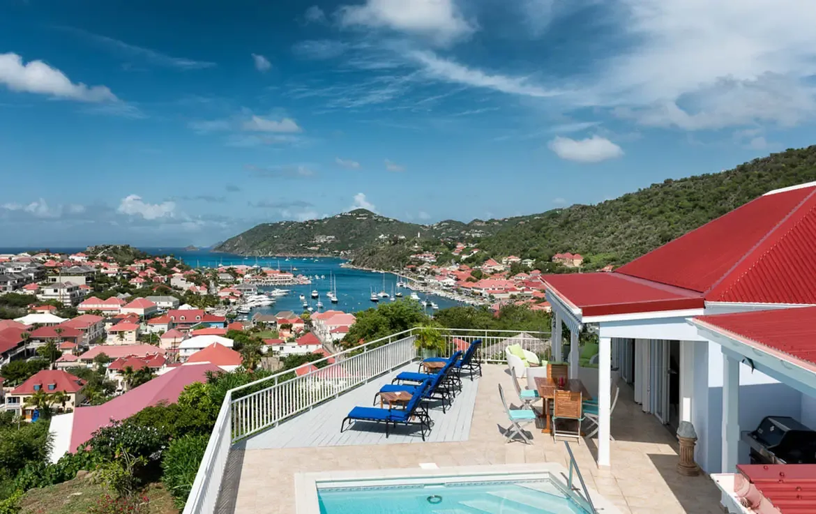 The expansive terrace frames a world-class harbor view where red rooftops pop against turquoise waters. Four plush loungers line the railing, a sparkling pool beckons below, and a covered veranda with outdoor kitchen awaits sunset cocktails—pure Caribbean living at its finest.