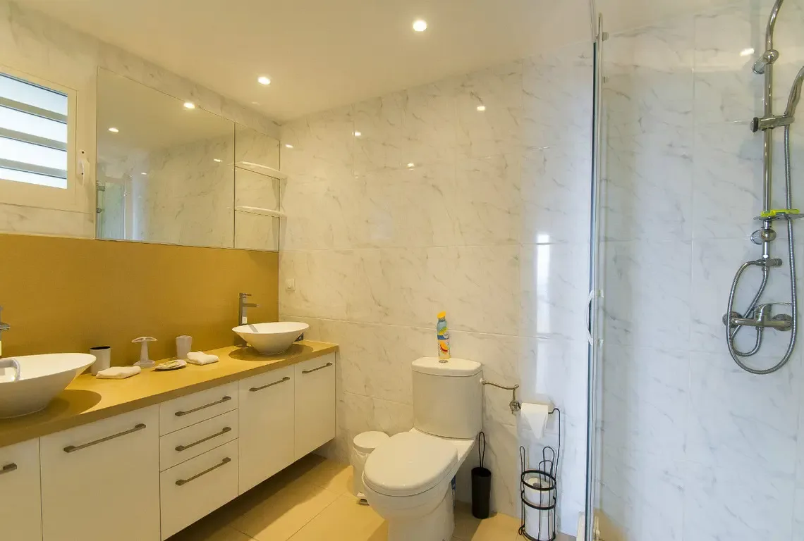 A contemporary bathroom with dual white vessel sinks set against a warm gold backdrop. The glass-enclosed walk-in shower features a rainfall showerhead, while marble-style walls and recessed lighting create a spa-like atmosphere.