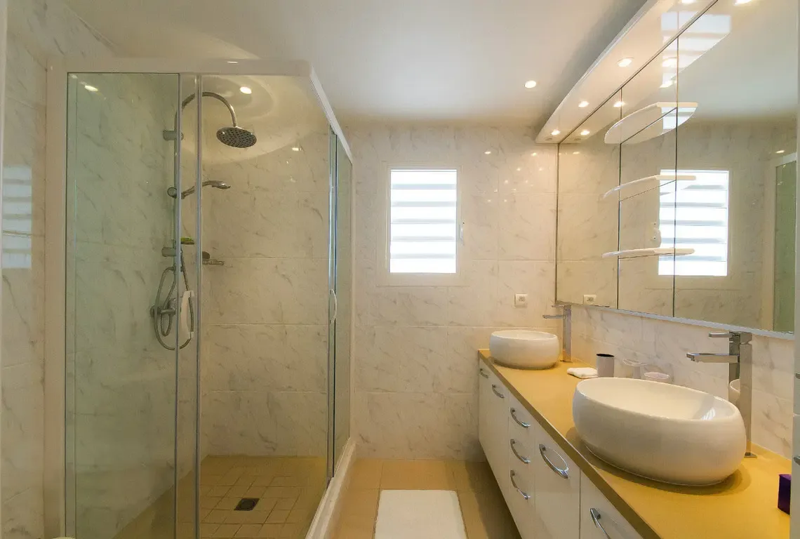 A spa-inspired master bathroom with seamless glass walk-in shower featuring rainfall showerhead and handheld wand. The double vanity with vessel sinks sits on a warm tan countertop, while marble-patterned walls and oversized mirrors create a serene retreat.