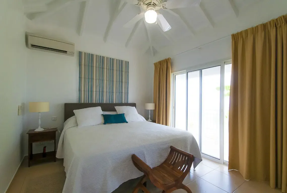A blue-and-white striped headboard panel adds coastal charm behind the king-sized bed with crisp white linens. Sliding glass doors flood the vaulted-ceiling room with natural light, while a sculptural wooden chair brings organic warmth to the serene space.