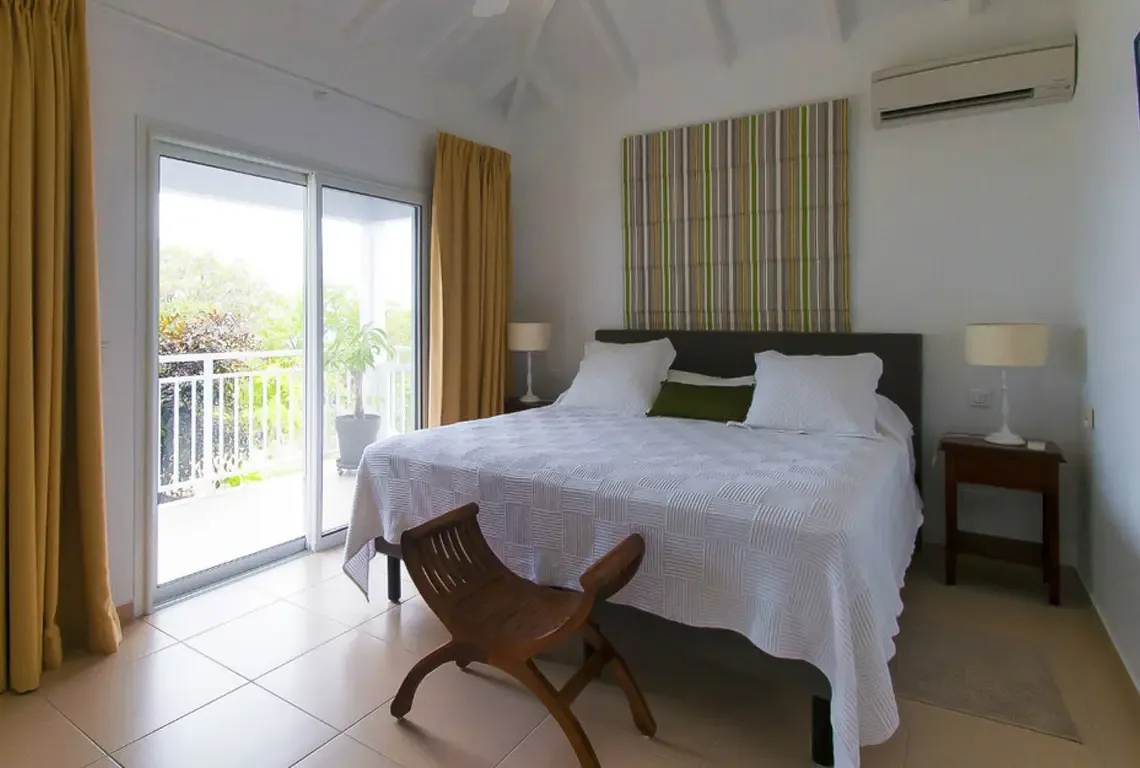 A king-sized bed with a striking striped headboard anchors this bright retreat. Mustard-yellow drapes add warmth while sliding glass doors open to lush tropical views. A curved wooden accent chair and vaulted ceilings complete this serene sanctuary.