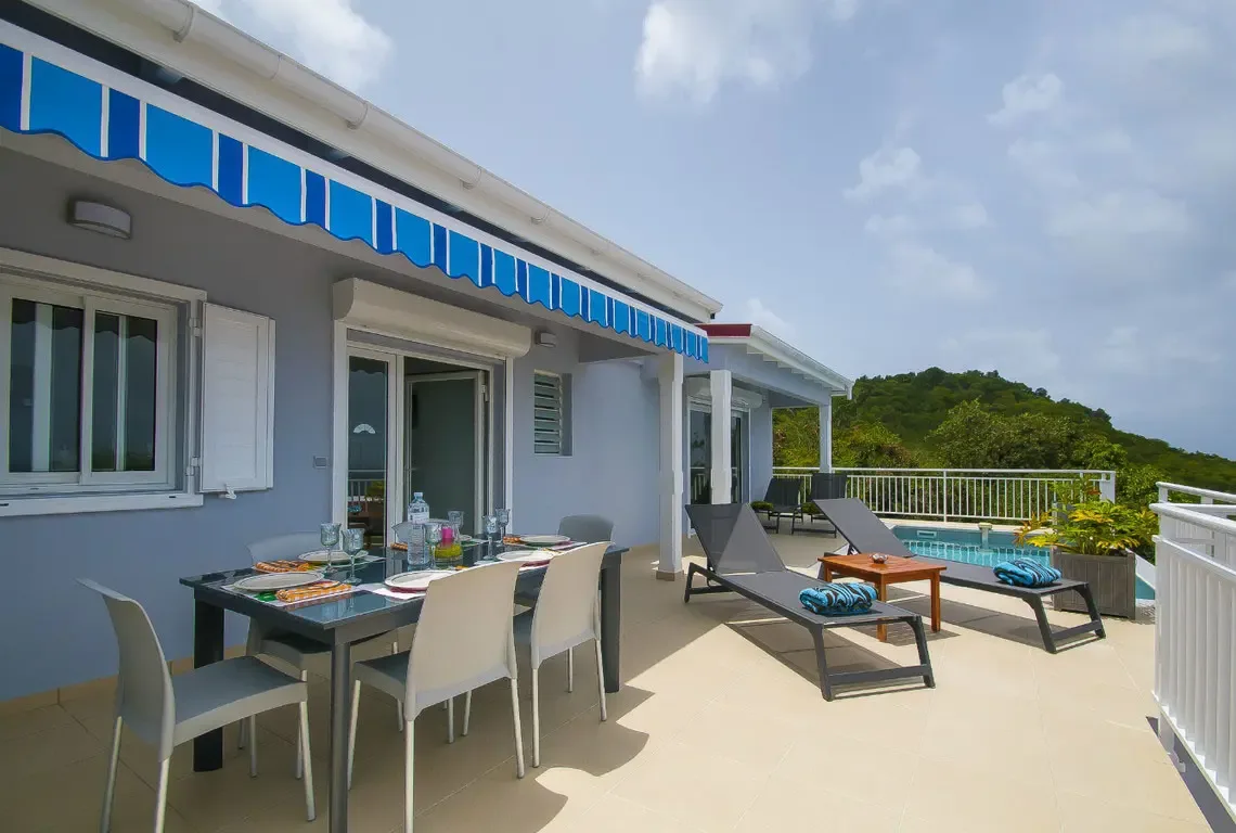 A blue-and-white striped awning shades the contemporary dining set while two loungers face the sparkling pool. Sliding glass doors connect to the interior, creating a seamless flow between cool indoor comfort and the sun-drenched terrace.