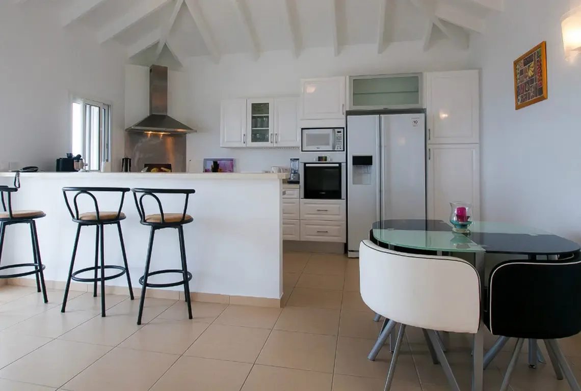 A professional stainless steel range hood crowns a rustic built-in oven. White cabinetry, a breakfast bar with three stools, and a glass-topped dining table fill this vaulted-ceiling space where cooking becomes a social affair.