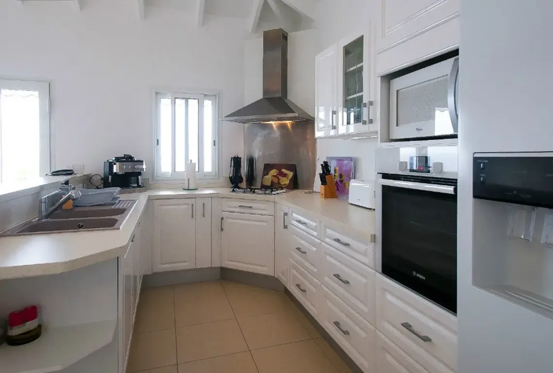 An all-white kitchen with expansive countertops and high-end stainless steel appliances, including a professional cooktop under a sleek range hood. Coffee maker, toaster, and a large refrigerator with water dispenser make mornings effortless.