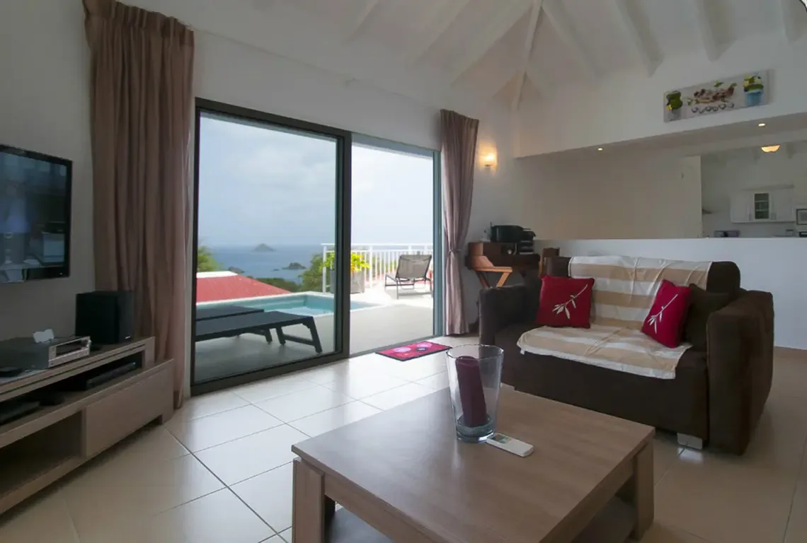 Floor-to-ceiling glass doors frame the turquoise sea while opening directly to your private pool terrace. A plush brown sofa with red accents faces the entertainment center, and the open-concept design flows effortlessly into the raised kitchen and dining area.