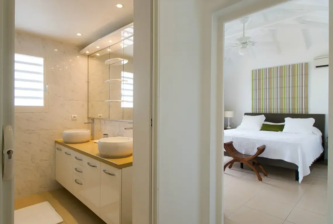 A seamless open-plan master suite where the spa-style bathroom flows into the bedroom retreat. White vessel sinks on warm wood, marble walls, and a king-sized bed with green accents create a cohesive, five-star sanctuary for ultimate relaxation.