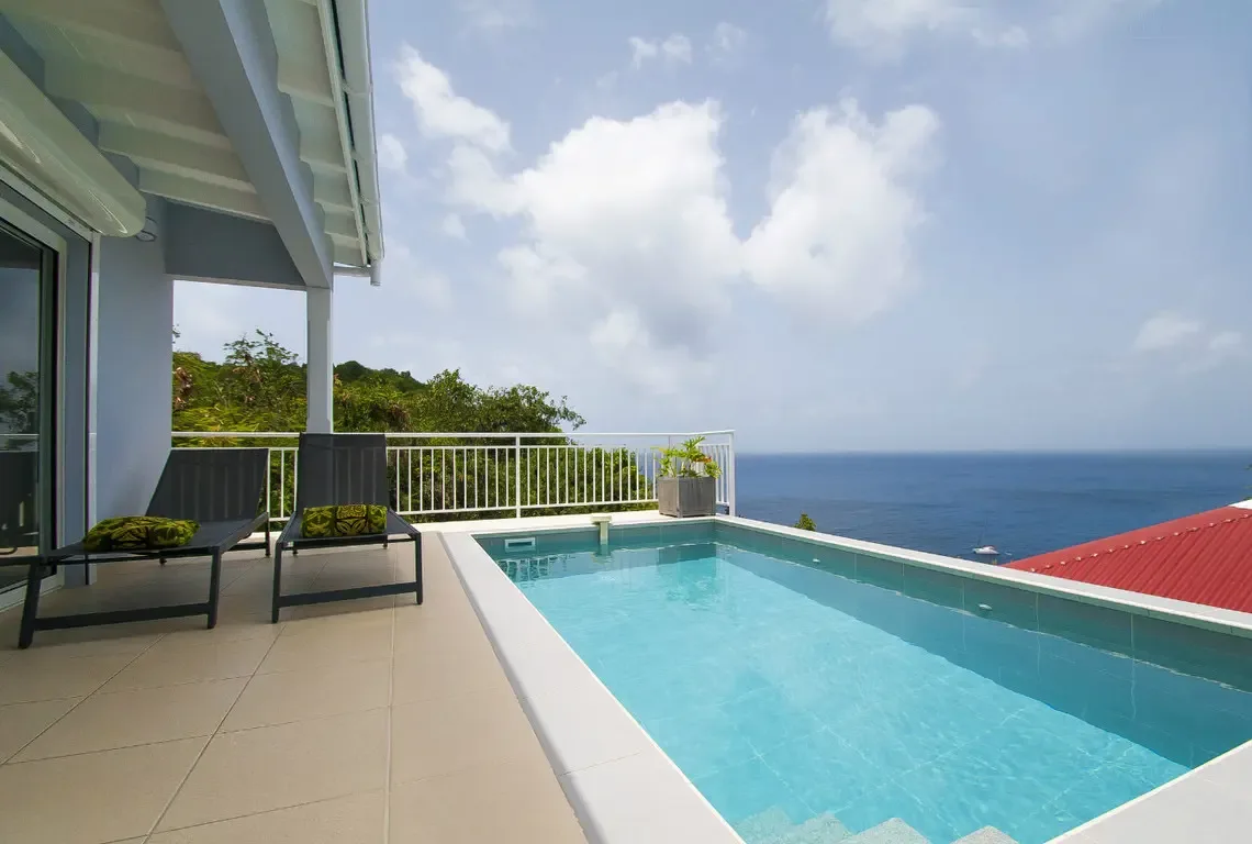 The infinity-edge pool seems to merge with the Caribbean horizon, while two sleek loungers with green pillows invite afternoon relaxation. Framed by tropical greenery, this elevated terrace delivers uninterrupted coastline views from a private hillside sanctuary.