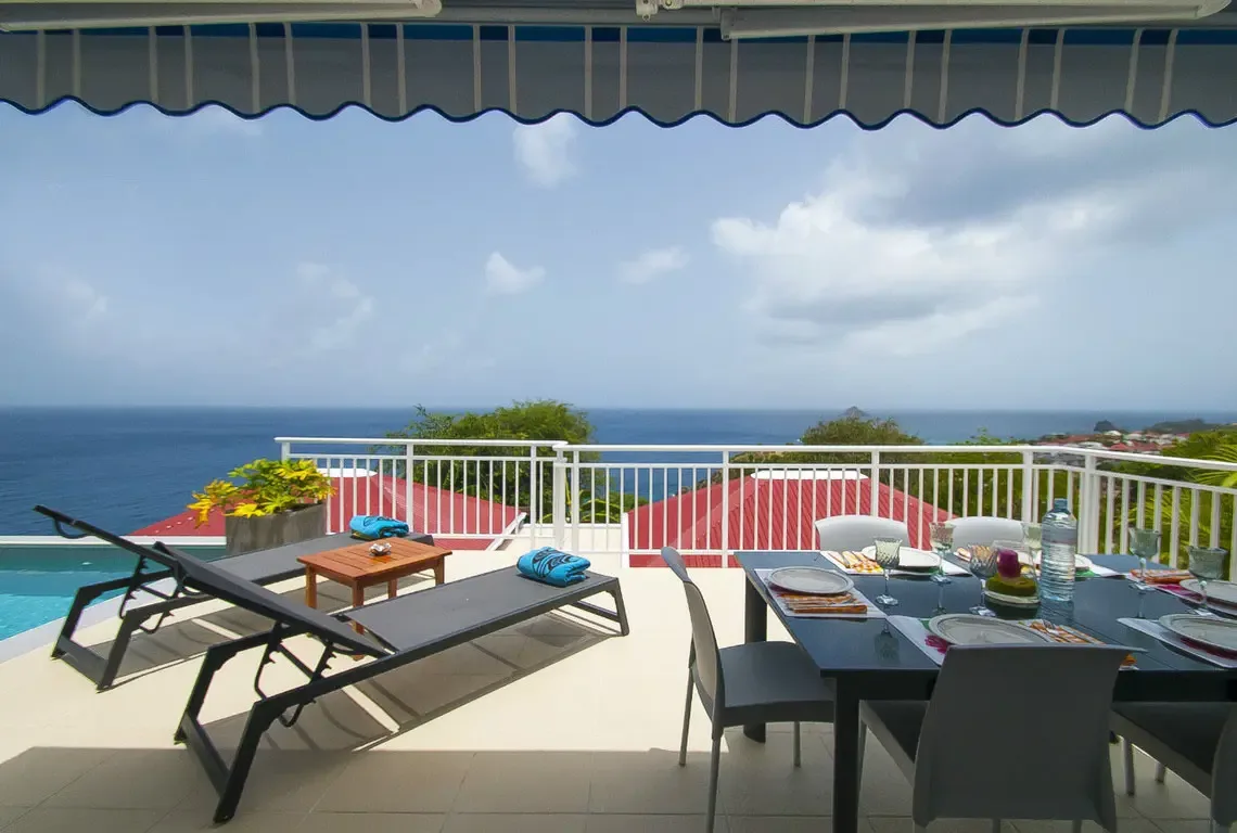 An elegant black dining table set for eight beneath a scalloped blue-and-white awning, with charcoal loungers and turquoise towels ready for sun-soaked afternoons. The infinity pool glimmers at the edge while red rooftops dot the lush hillside below.