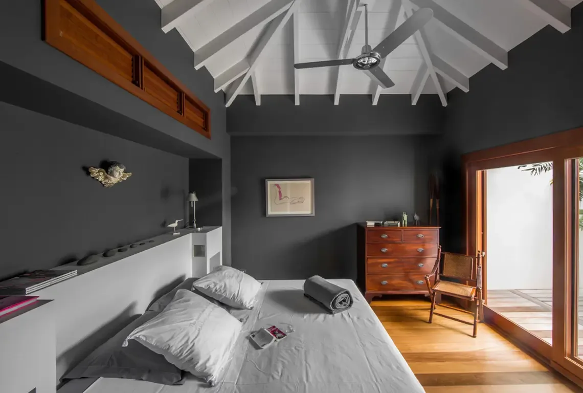 A minimalist bedroom with dark grey walls and white vaulted beamed ceiling. A built-in headboard shelf displays curated objects—magazines, stones, a bird figurine. Sliding glass doors open to the outdoor patio while a silver ceiling fan keeps the space cool.