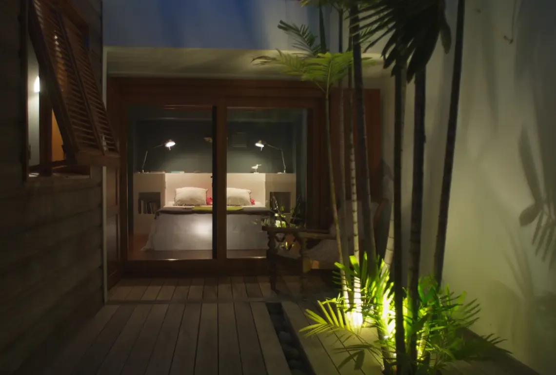 A nighttime view into a minimalist bedroom with two twin beds against a dark grey wall. Warm spotlights illuminate palm plants on the wooden deck outside, while modern adjustable lamps provide reading light—a tranquil, private retreat after sunset.