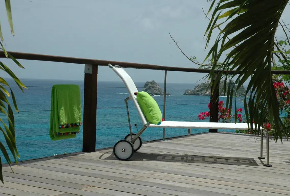 A sleek white chaise lounge with lime-green pillow faces the turquoise Caribbean through stainless-steel cable railings. Weathered hardwood deck, swaying palms, and red bougainvillea frame your private sanctuary—uninterrupted relaxation with iconic rocky islets on the horizon.