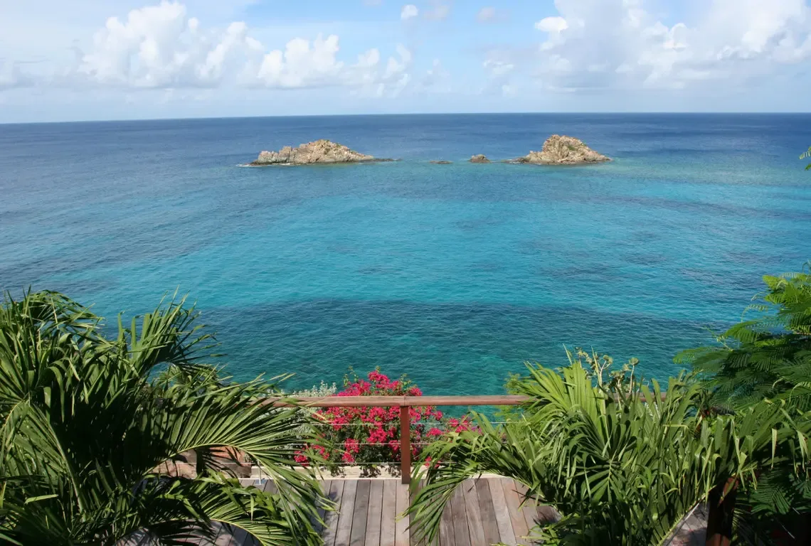 From your weathered wood deck, the Caribbean stretches to the horizon in shades of sapphire to translucent turquoise. Pelican Rocks rises from the sea while pink bougainvillea and swaying palms frame the view—your private perch above paradise.