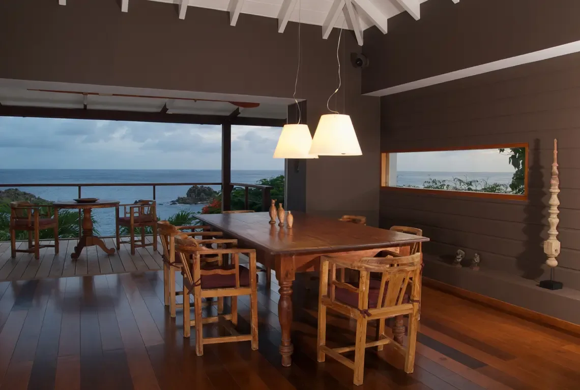 A dining area with four chairs surrounds a wooden table beneath pendant lights, with brown walls and white exposed beams. Through the opening, the sea and outdoor deck are visible—indoor dining meets Caribbean panorama.