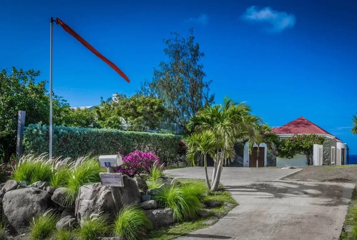A vibrant red windsock catches the trade winds above this prestigious Gustavia Hill entrance. Manicured hedges, weathered boulders, and magenta bougainvillea frame the classic red-tiled roof and arched doorways—your gateway to a private hilltop retreat.