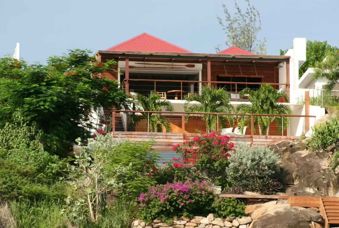 Red corrugated roofs crown this hillside sanctuary where warm wood siding meets natural stone. Multiple deck levels cascade down the slope, while open-air portals on the upper floor invite Caribbean breezes—your private jungle paradise awaits.