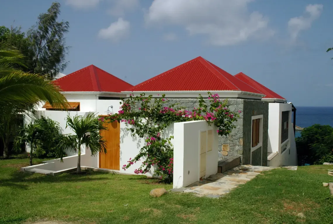 Steep red corrugated roofs crown this hillside retreat, contrasting beautifully against white masonry and natural stonework. Pink bougainvillea cascades over the walls while palm fronds sway—bold modern architecture meets Caribbean tranquility.