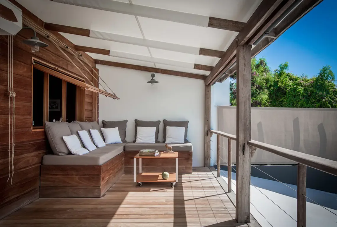 A dark wood sofa with grey and white cushions anchors this indoor-outdoor living space. A wheeled table with books and a white sculpture sits center stage, while sunlight streams through the open porch where a white deck meets lush tropical greenery.