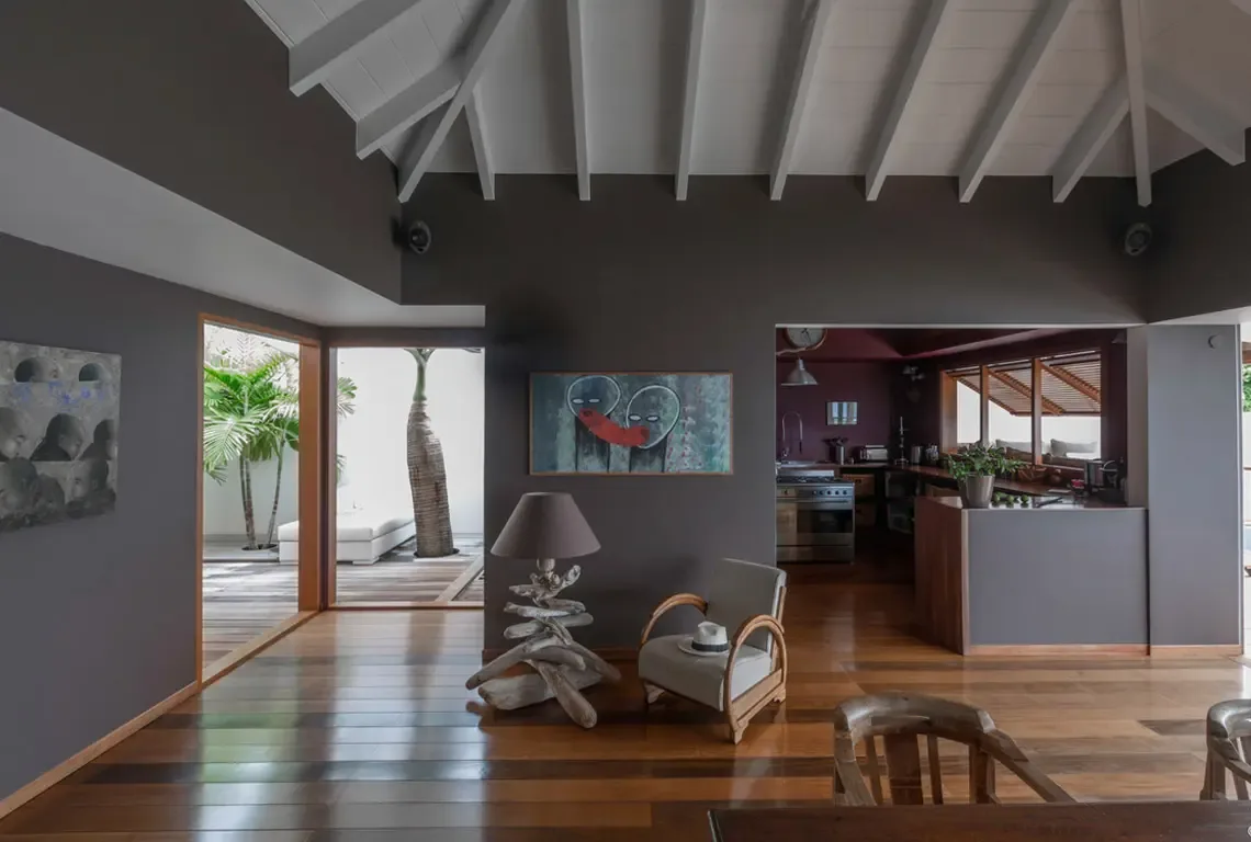 Vaulted white rafters crown this dramatic living space where charcoal walls meet polished hardwood floors. A contemporary painting, driftwood floor lamp, and rattan armchair with white fedora create curated sophistication—glass doors open directly to the tropical deck beyond.