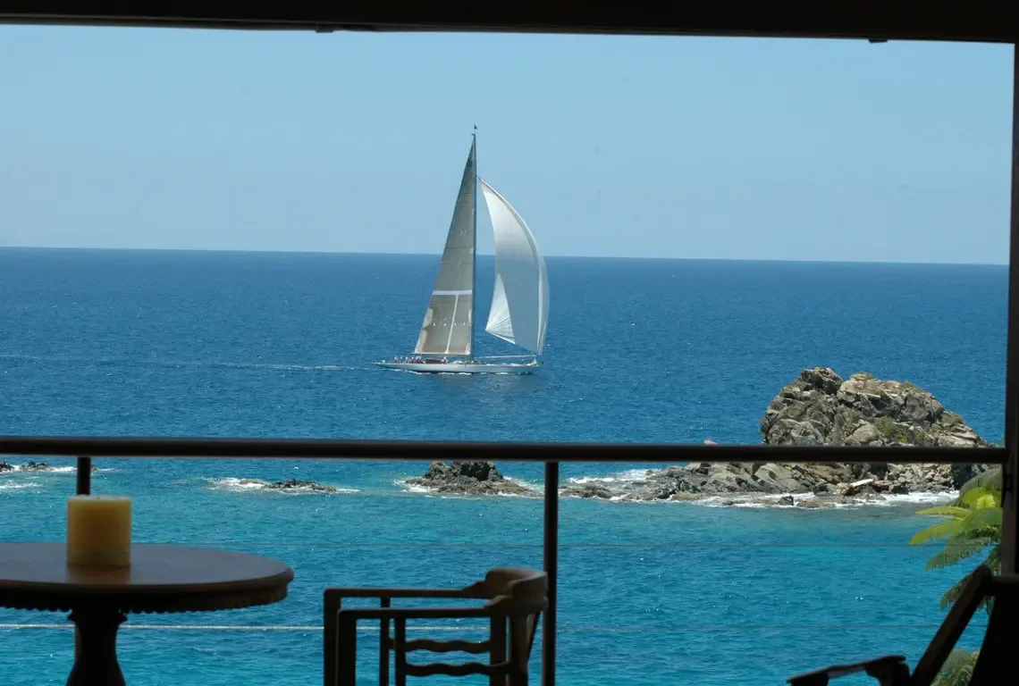 A sailing yacht glides across the bay, framed perfectly by the balcony's picture-window view. Inside, a round wooden table with pillar candle and comfortable chairs bridge indoor comfort with the nautical scene—your front-row seat to maritime elegance.