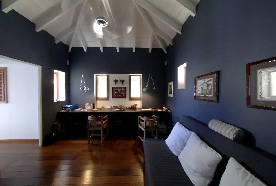 A second angle of the study showcases dark blue walls and warm wood floors beneath white-beamed ceilings. Multiple windows frame tropical views while artwork adorns the walls—natural light filters through louvered shutters, creating soft shadows and an inspiring atmosphere.