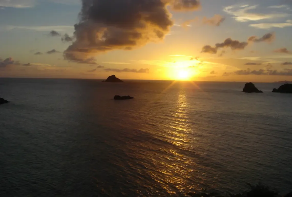 The sun dips toward the horizon, painting the sky in gold, amber, and peach. A shimmering path of light reflects across the tranquil sea while rocky silhouettes frame your private sunset ritual—nature's daily masterpiece from your hillside perch.