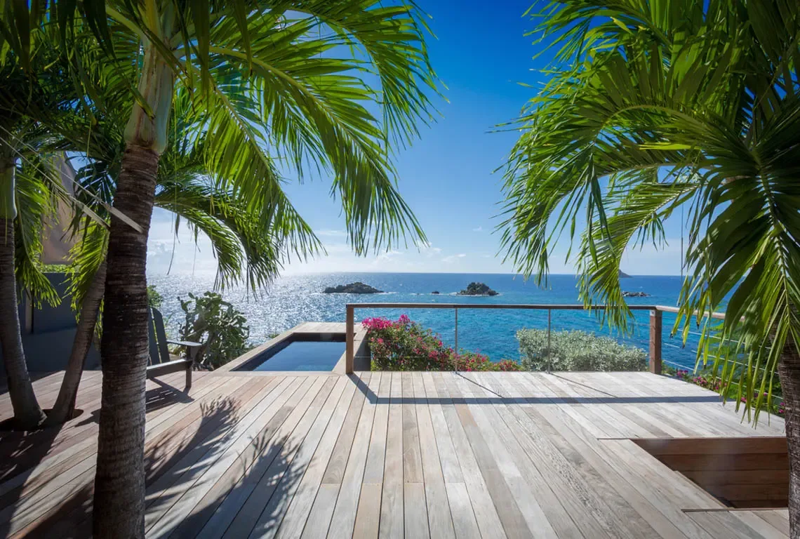 A weathered wood deck with integrated plunge pool faces the turquoise Caribbean, framed by swaying palms and vibrant bougainvillea. Glass railings keep the panoramic view unobstructed—your private front-row seat to Caribbean beauty.