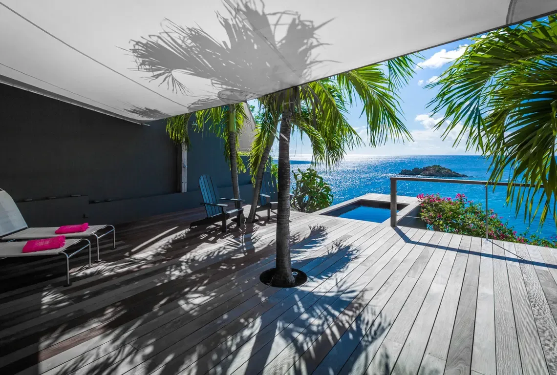 A mature palm grows through a deck cutout beneath a white canopy that casts dappled shadows. The square plunge pool merges with the turquoise horizon while pink towels brighten sleek loungers—tropical chic at its finest.