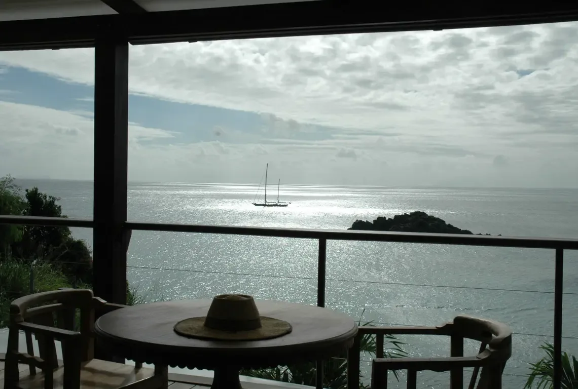 A covered veranda with dark-wood armchairs frames the sparkling bay where sunlight dances on the water. A straw hat rests on the table, a sailboat anchors in the distance—your shaded sanctuary for slow mornings and peaceful afternoons.