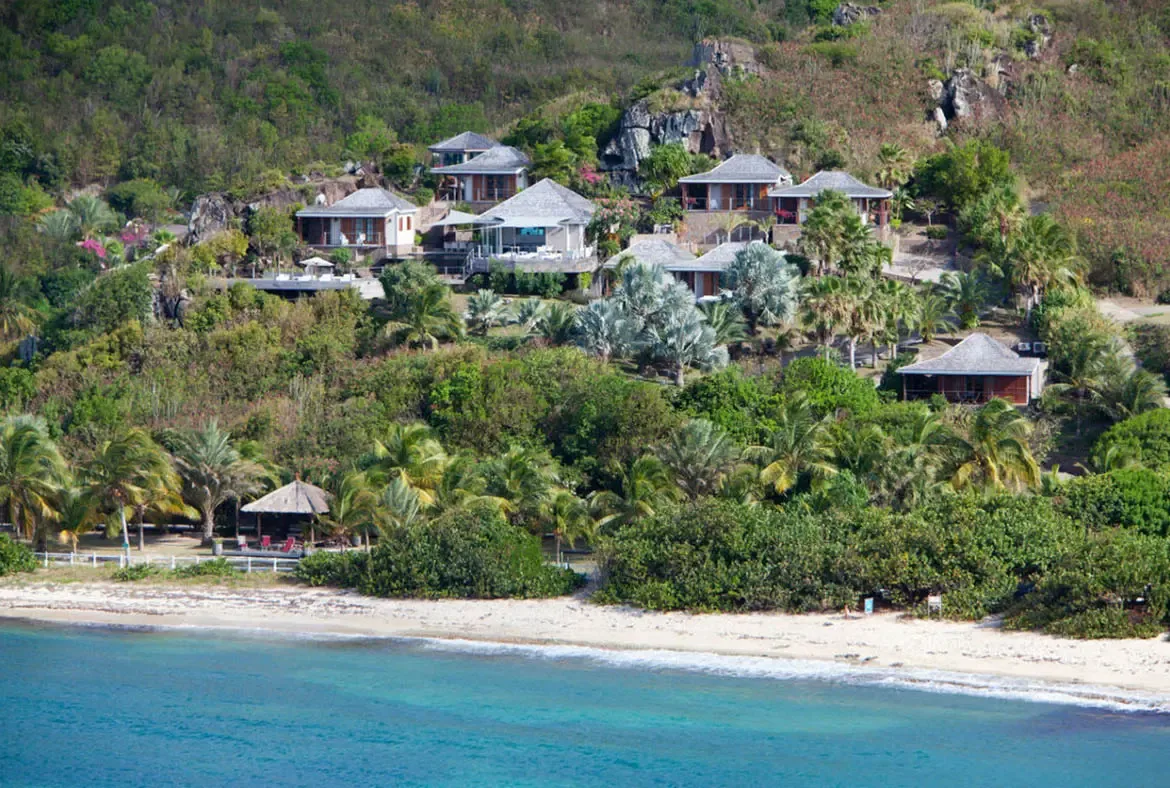 An aerial view captures a terraced villa compound with grey-shingled roofs nestled into a lush hillside, where a thatched beach gazebo and turquoise water complete a secluded paradise.
