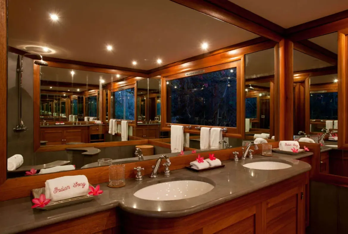 A double stone vanity with chrome faucets and white oval sinks stretches under wall-to-wall mirrors, where pink frangipani blossoms and "Indian Song" monogrammed towels add tropical elegance.