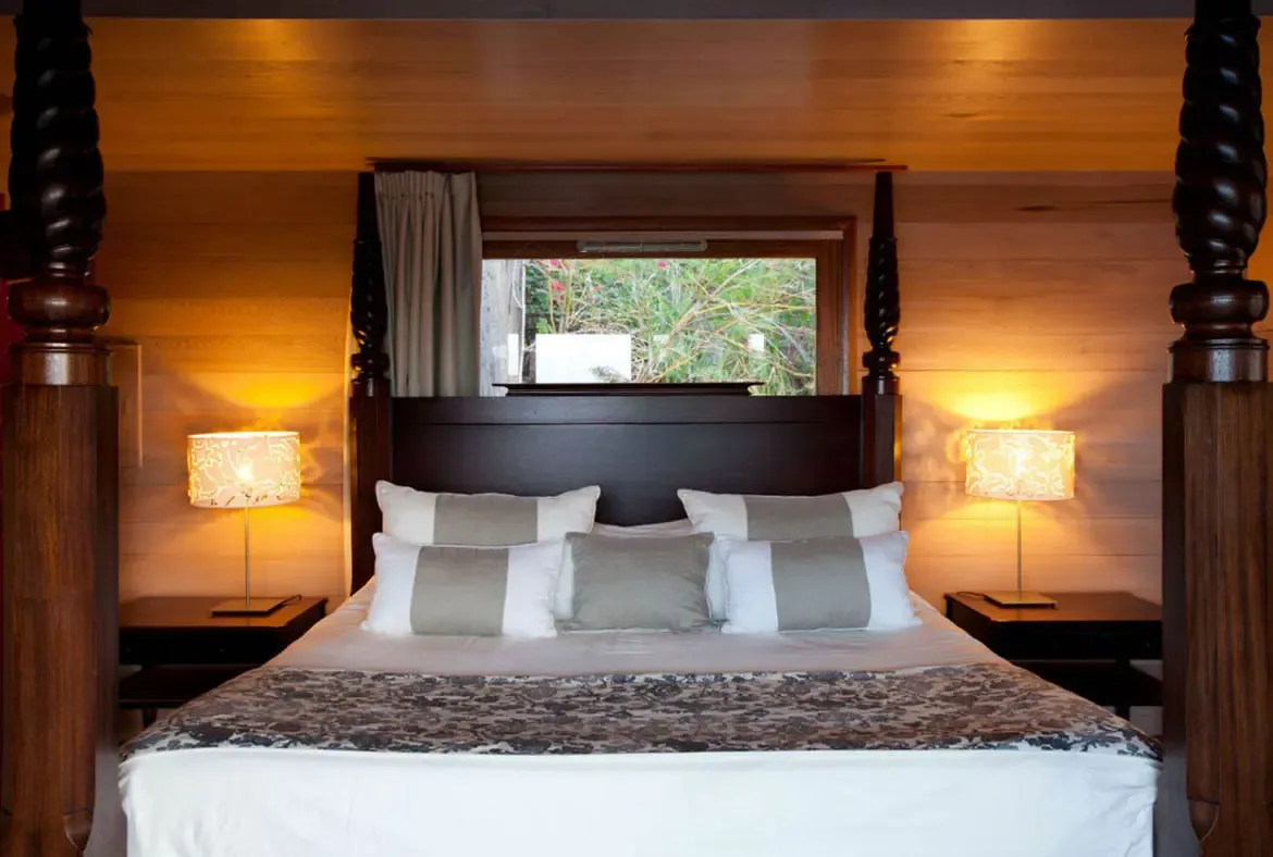 A dark carved four-poster bed with white linens and a grey floral runner sits in warm wood-paneled walls, where matching bedside lamps cast a golden glow across the cozy bedroom.
