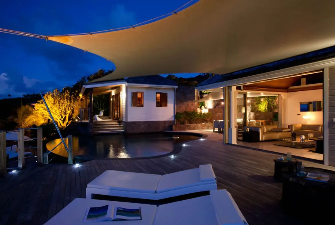 White loungers and an open book rest on a dark deck under a sail shade, where open glass doors reveal a warm living room and a curved infinity pool meets twilight.