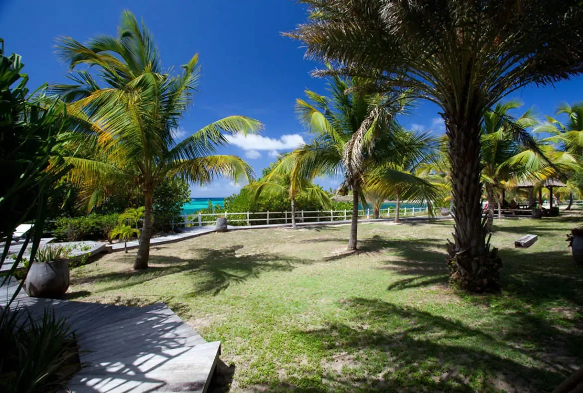 Mature palms cast long shadows across a green lawn from a wooden walkway, where a white fence and a thatched gazebo overlook turquoise ocean under a bright blue sky.