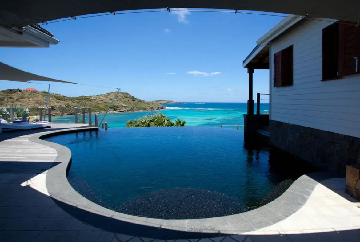 A dark-tiled infinity pool with a curved edge mirrors the sky above, where turquoise shallows reveal coral reefs and green hills curve around a pristine Caribbean bay.