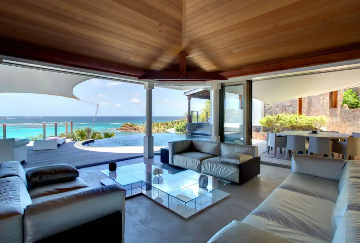 Silver-grey sofas and a glass-block coffee table sit under a warm wood ceiling, where open glass doors lead to a curved infinity pool and turquoise bay stretching to rocky islands.