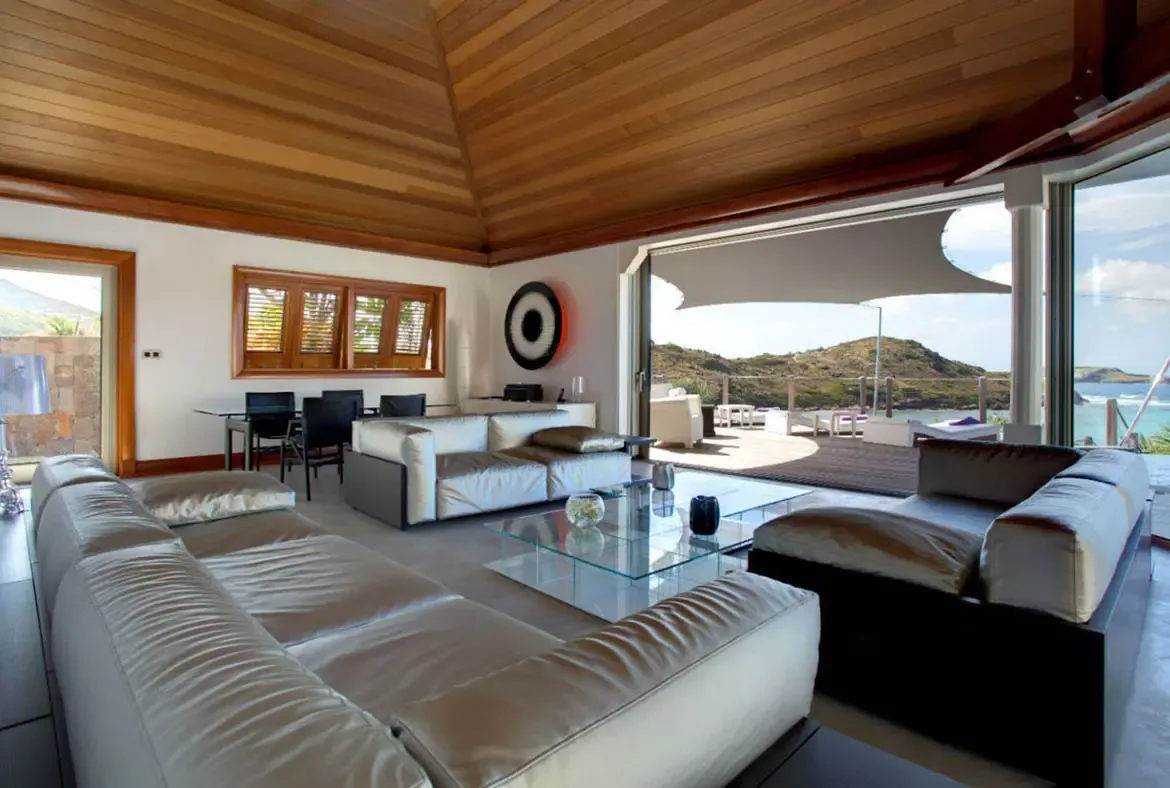White leather sofas and a glass coffee table sit under a vaulted wood ceiling, where open doors reveal a sail-shaded terrace and turquoise ocean beyond a rocky coastline.