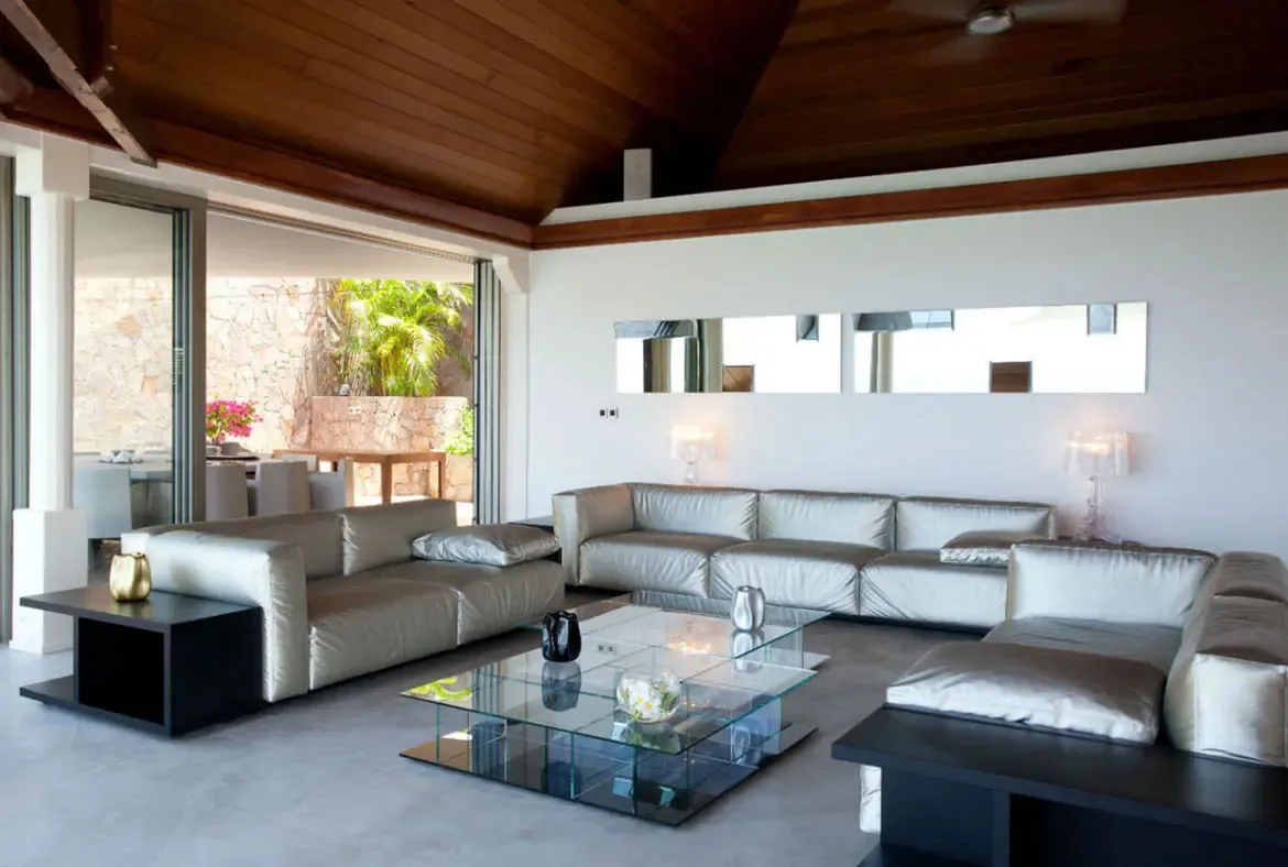 U-shaped silver sofas and a glass-block coffee table face a mirror wall and target art, where open glass doors reveal pink bougainvillea and a sun-drenched terrace.
