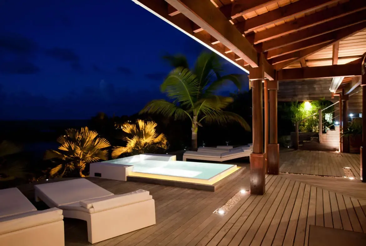 A glowing white plunge pool sits on a dark wooden deck under a gabled roof, where uplit palms and recessed floor lights create a cinematic nighttime retreat.