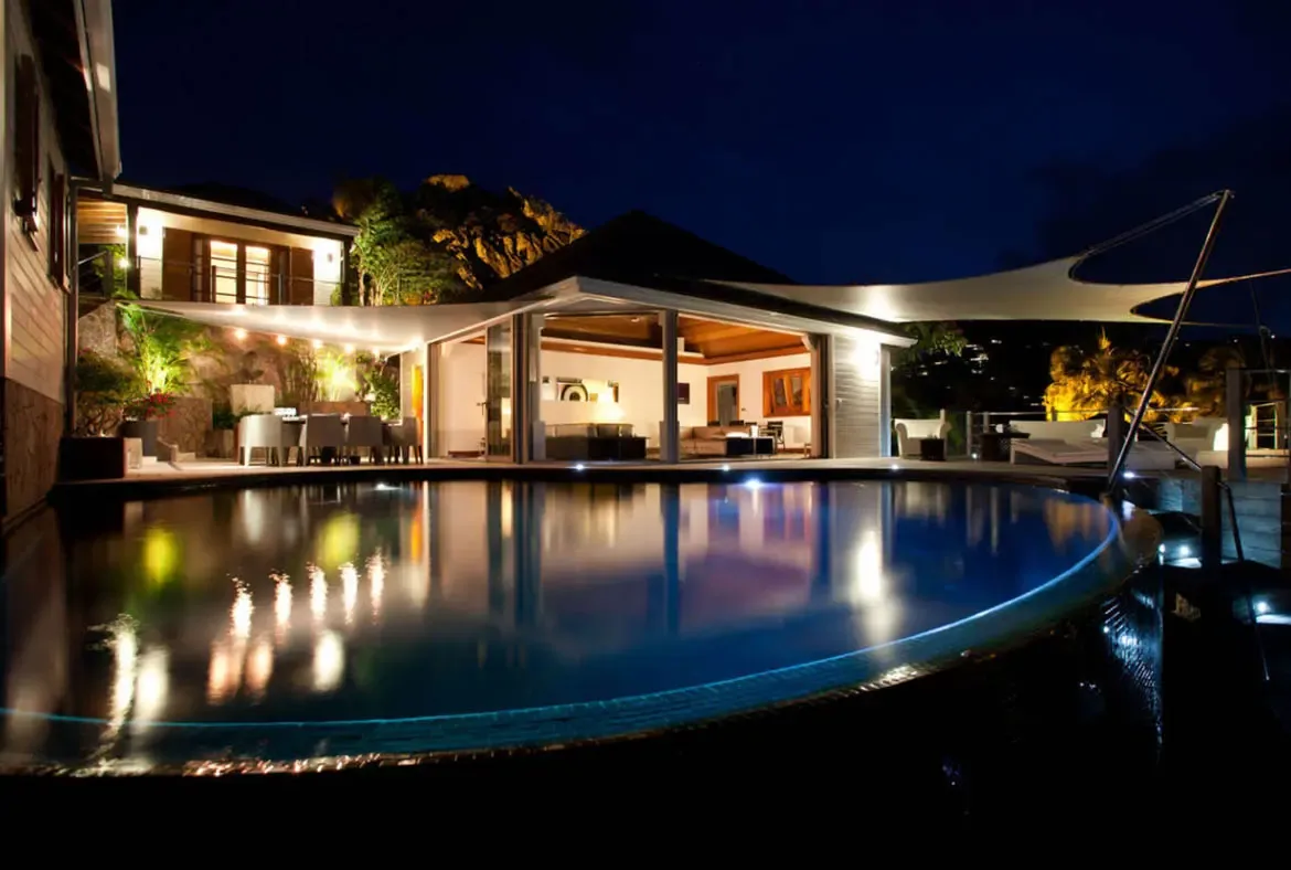 At night, a still curved pool mirrors golden villa light and white sail canopies, where open glass doors and an illuminated deck create a cinematic indoor-outdoor retreat.
