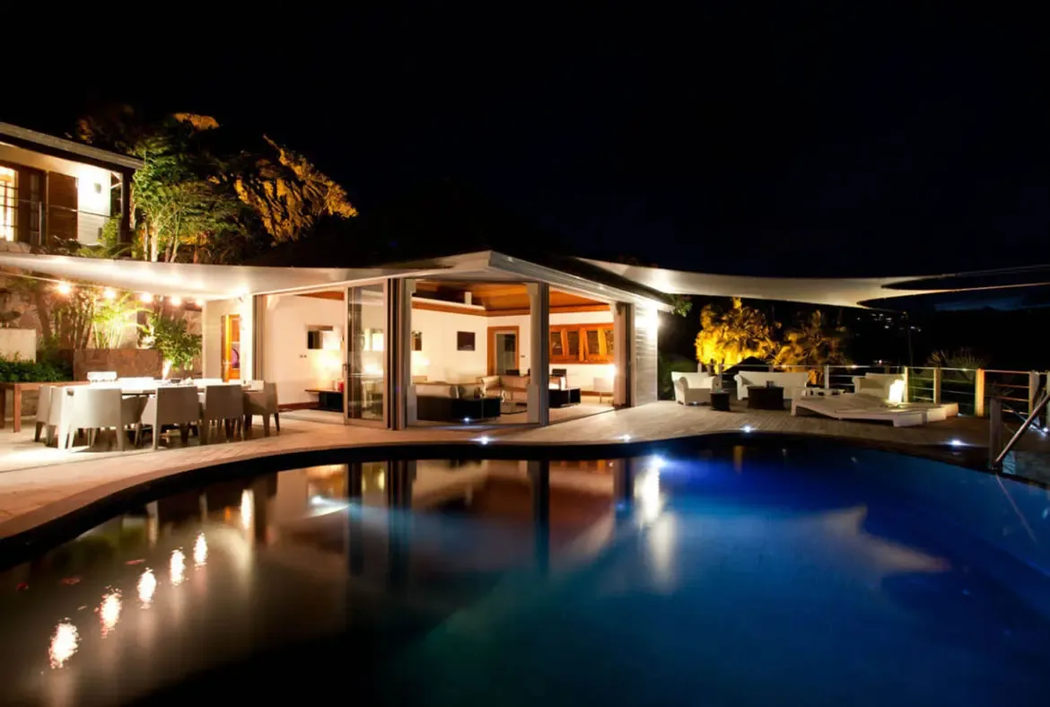 Warm light glows from open living spaces under white triangular sail shades, where a dark curved pool mirrors the villa's golden interior against a deep blue night sky.