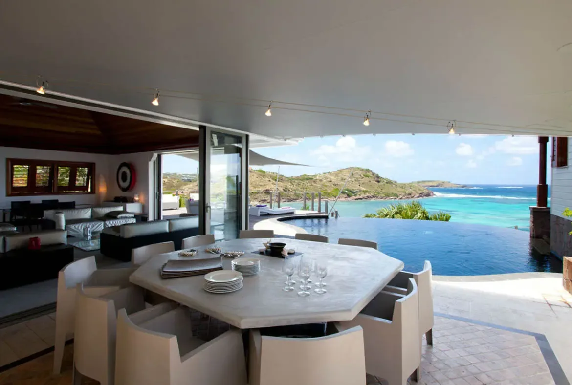 A stone dining table set with white plates and wine glasses sits under a white overhang, where a dark infinity pool merges with turquoise ocean and green hills beyond.